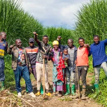 A group of people stand in a field surrounded by tall green crops, some are holding farming tools. They joyfully smile and hold up their thumbs.