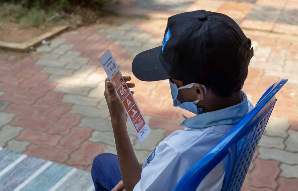 A young schoolboy, wearing a blue cap with a UNDP logo and a face mask, is seated outdoors attentively examining a pamphlet featuring diagrams and pictures. The setting appears to be educational or informational, possibly part of a disaster risk reduction community awareness program. Photo credits: UNDP Bangkok Regional Hub.