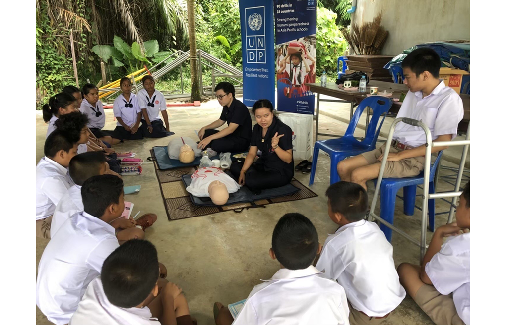 A group of schoolchildren, attentively seated on the ground, receive first aid training from a trainer. There's also a student with a walker, seated in a chair, listening to the training. A UNDP banner stands in the background. The setting is informal, under a shelter, which suggests a focus on practical, hands-on learning. Photo credits: UNDP Thailand.