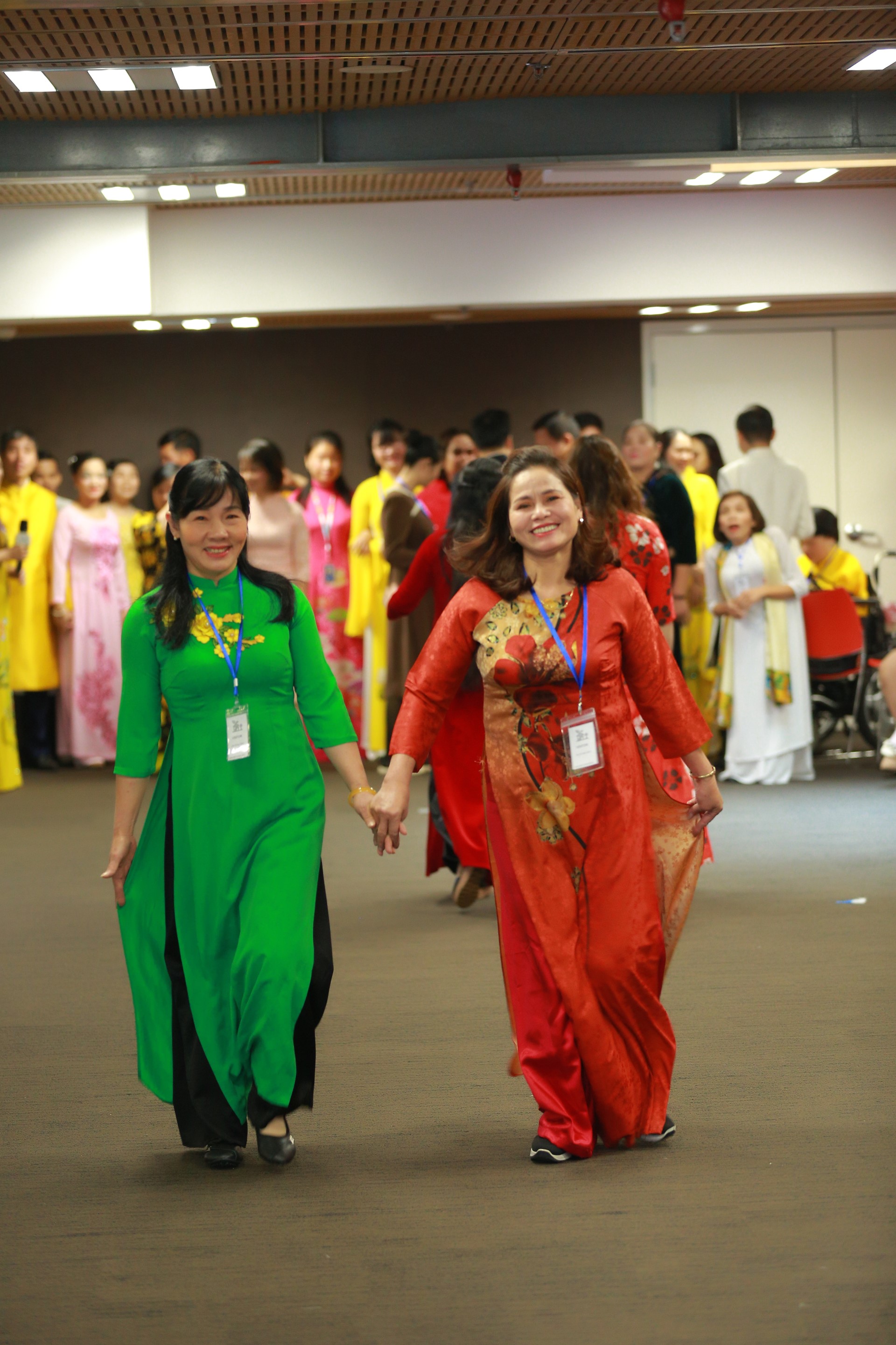 Two women in traditional Áo Dài dresses, one green and one red, walk hand in hand with smiles, amongst a group of people in colorful attire at an indoor fashion show. Photo credits: UNDP Vietnam. 